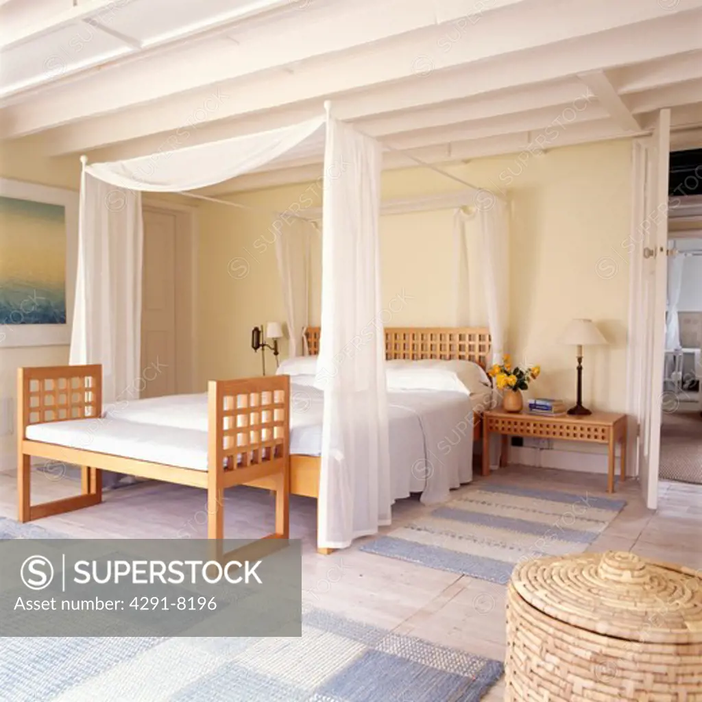 White voile curtains and linen on modern four poster bed with matching stool in cream bedroom with white ceiling beams