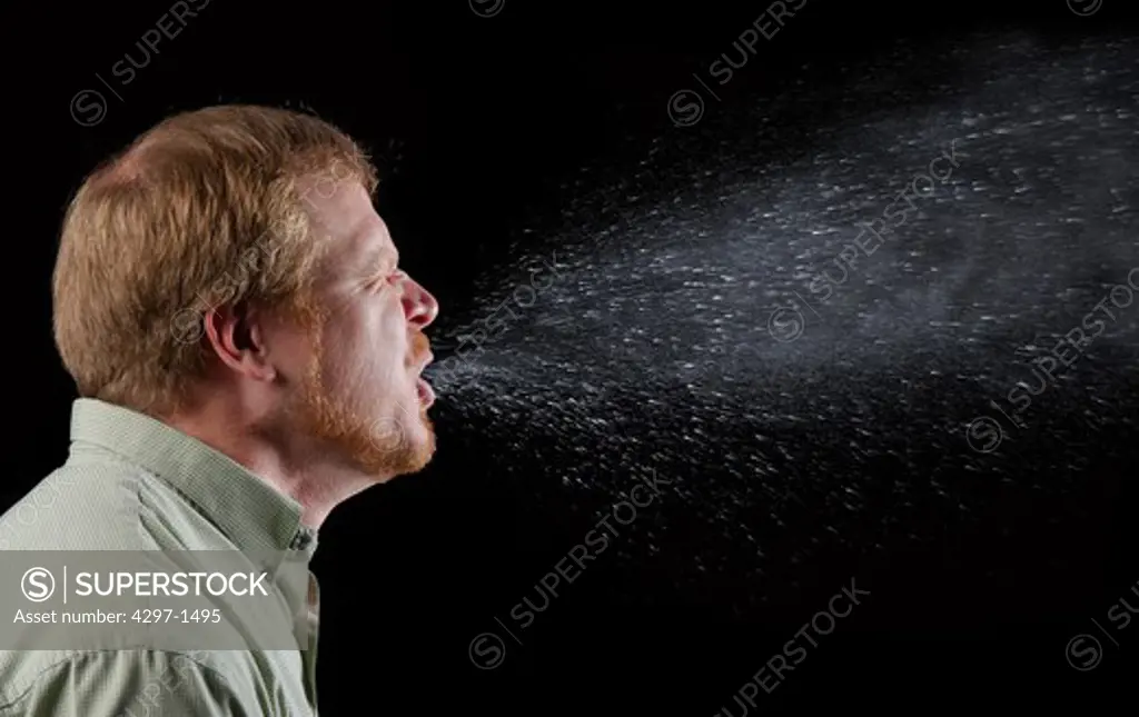 Photograph capturing a sneeze in progress, revealing the plume of salivary droplets as they are expelled in a large cone-shaped array from this man's open mouth