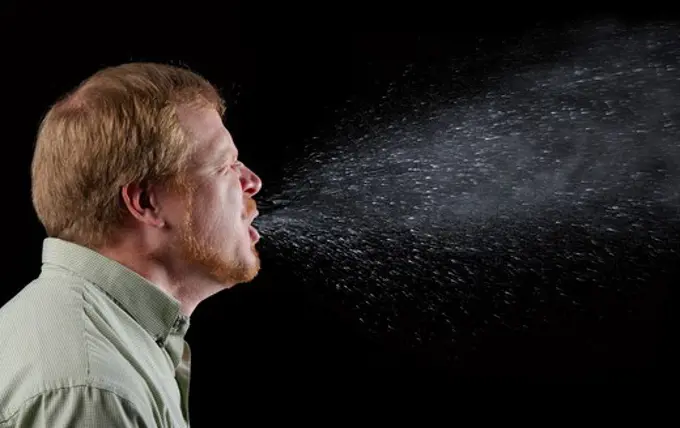 Photograph capturing a sneeze in progress, revealing the plume of salivary droplets as they are expelled in a large cone-shaped array from this man's open mouth