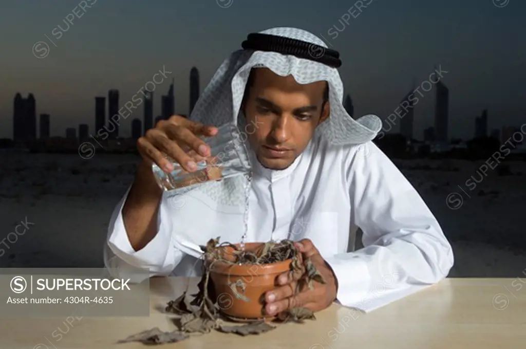 Young Arab man sitting at a desk with Dubai City in the background pouring water to a dead plant