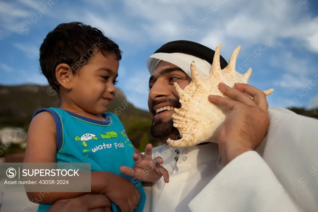 Arab father and son at the beach, man holding seashell to his ear.