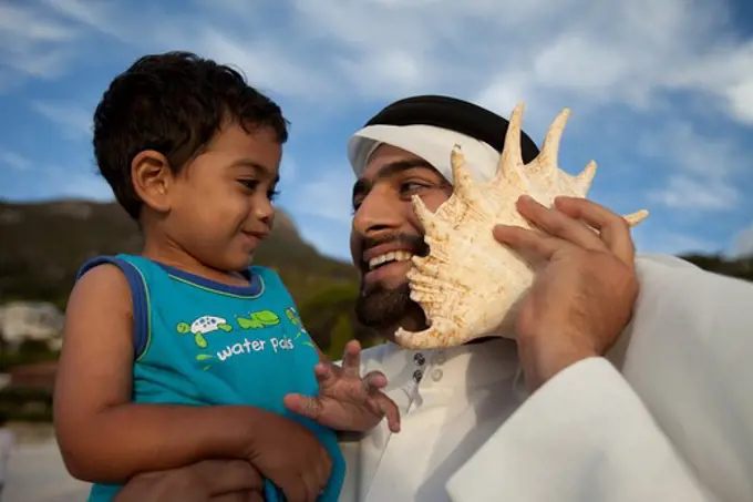 Arab father and son at the beach, man holding seashell to his ear.