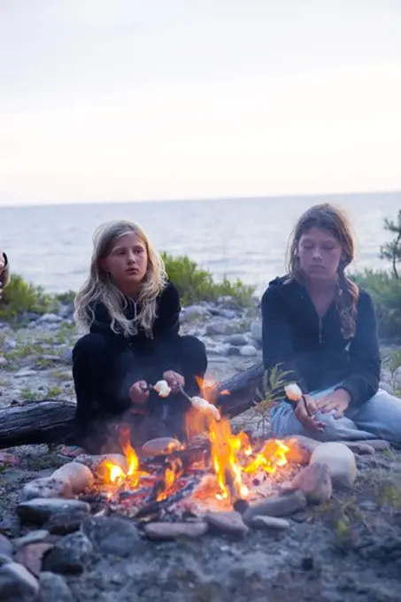 Two Scandinavian girls sitting by a fire barbequeing, Oland, Sweden.