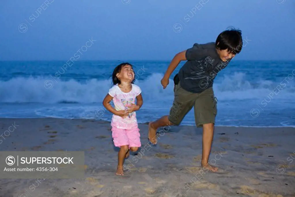 A brother and sister play on the beach at twilight.