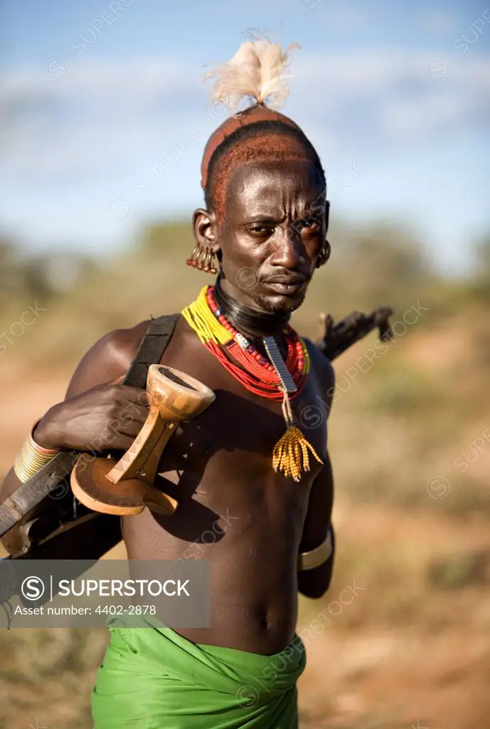 Hamar tribesman with feather headdress, carrying his rifle and a traditional wooden stool. Omo Delta, Ethiopia, Africa