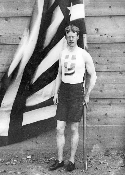 Athens (Greece), 04/10/1896. The American athlete Thomas Pelham Curtis poses next to his country's flag at the Olympic Games, where he will win the gold medal in the one hundred and ten meter hurdles.