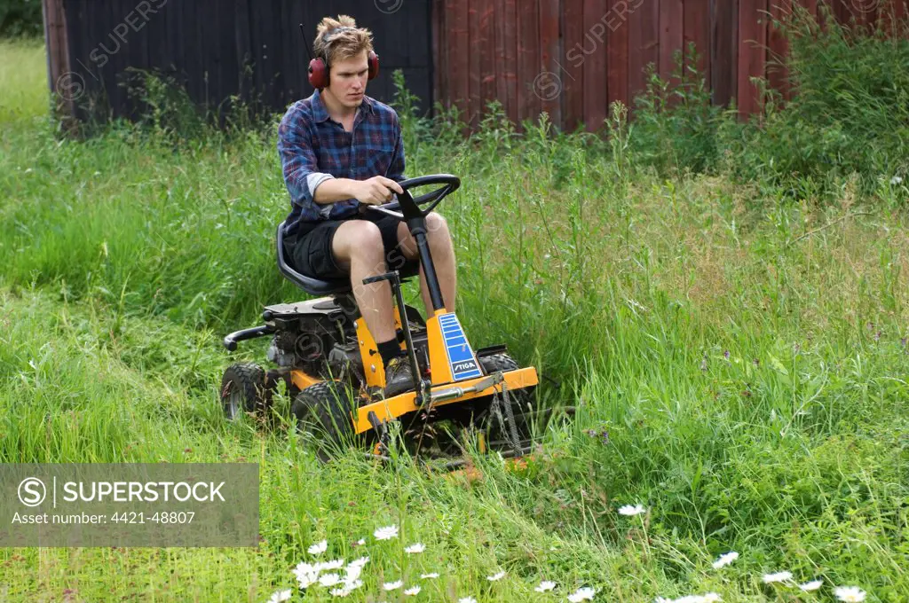 Man using ride-on mower on farm, cutting long grass around sheds, Sweden