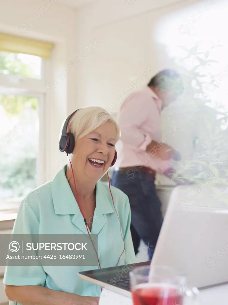 Happy senior woman with headphones video chatting at laptop in kitchen