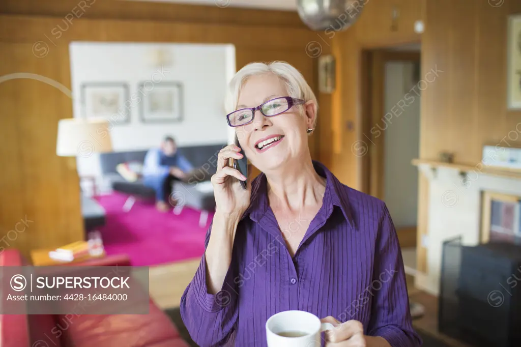 Happy senior woman with coffee talking on smart phone in living room