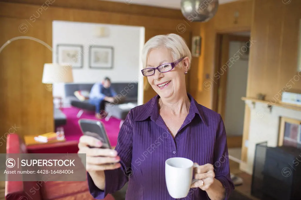 Happy senior woman using smart phone and drinking tea in living room