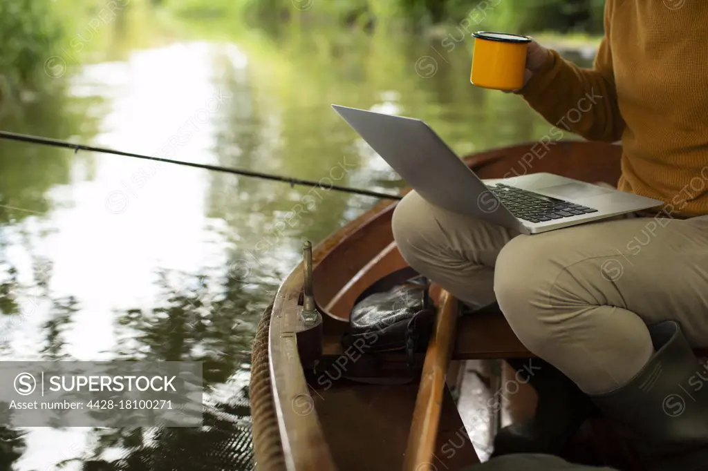 Man fly fishing and using laptop with coffee in boat on river