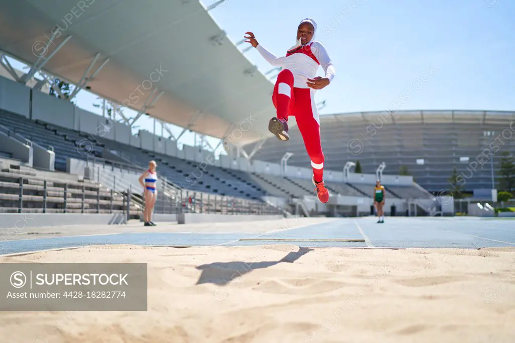 Female track and field athlete long jumping over sand in sunny stadium