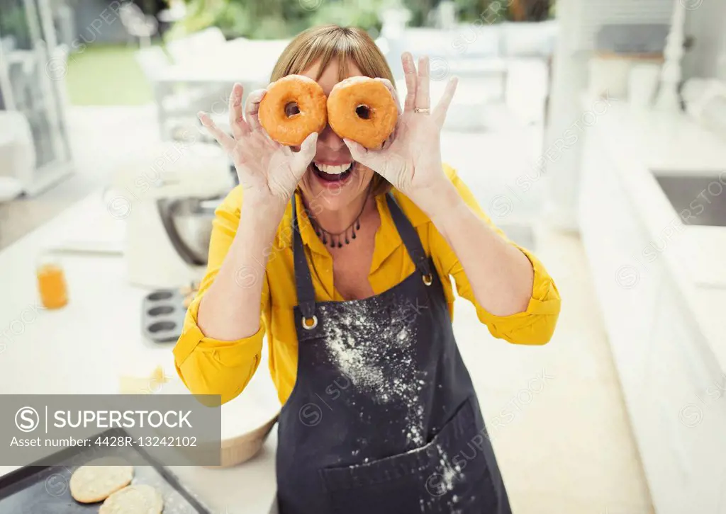 Portrait playful mature woman covering eyes with donuts in kitchen