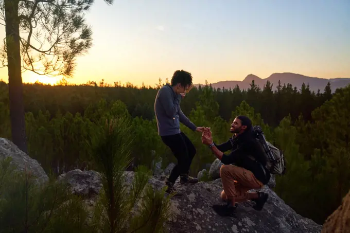 Young man with engagement ring proposing to woman on rock at sunset