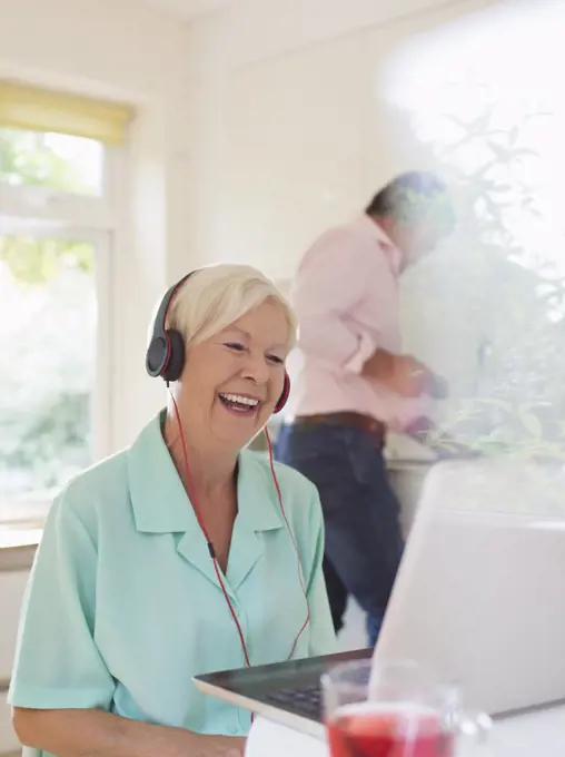 Happy senior woman with headphones video chatting at laptop in kitchen