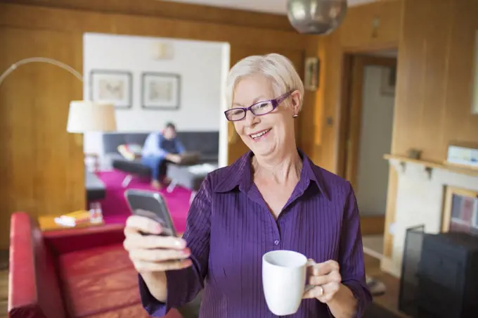 Happy senior woman using smart phone and drinking tea in living room