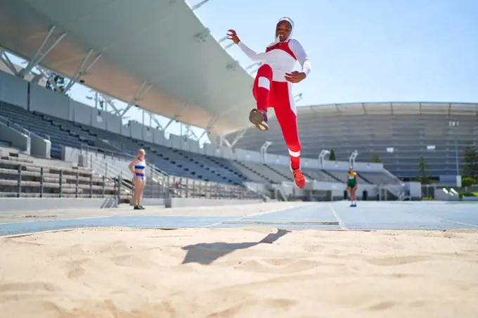 Female track and field athlete long jumping over sand in sunny stadium