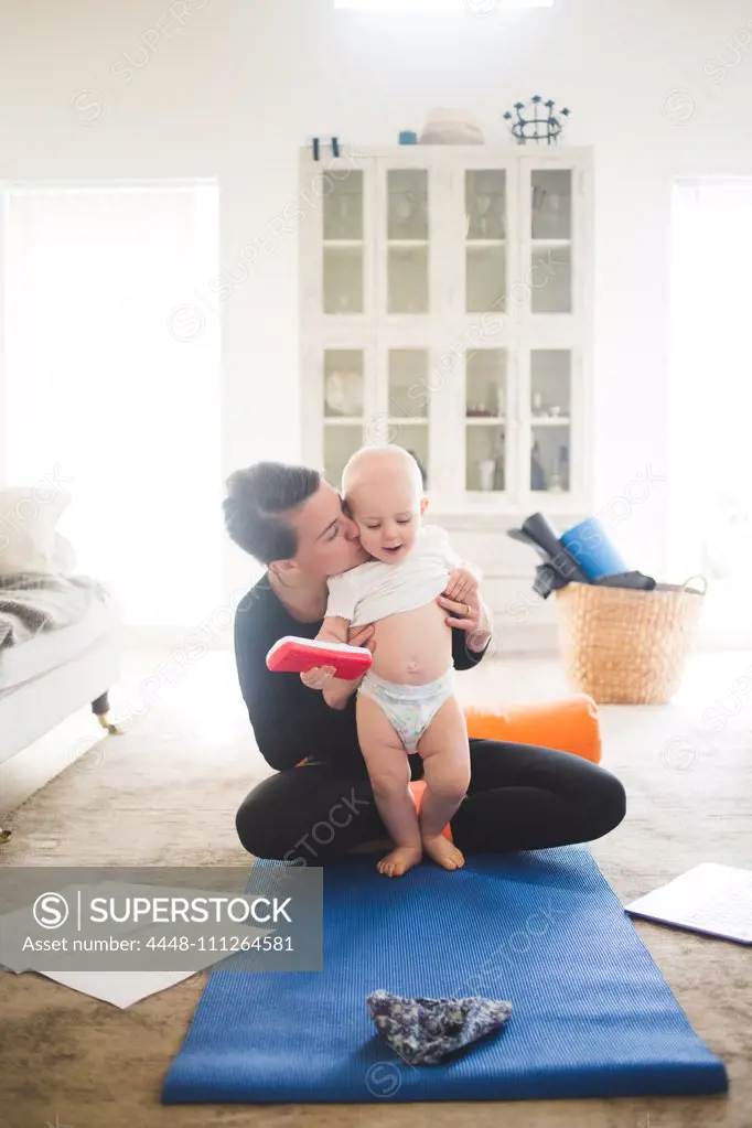 Female entrepreneur kissing daughter while practicing yoga in living room at home