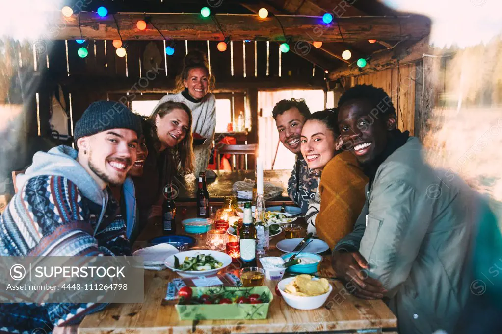 Portrait of smiling friends with food and drink on table in cottage seen through window