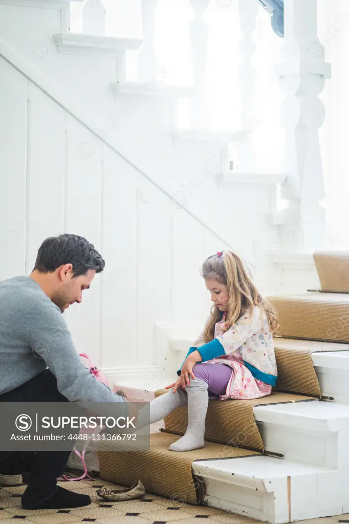 Father assisting schoolgirl wearing shoes while sitting on steps at home