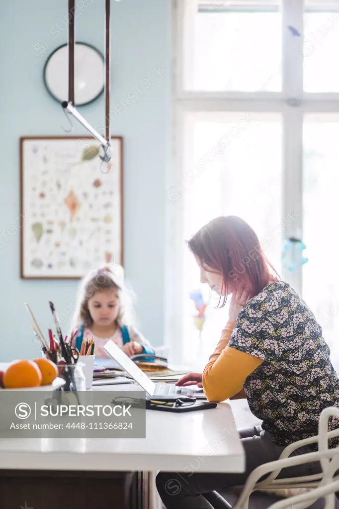 Side view of mid adult women working on laptop while sitting with girl at kitchen island