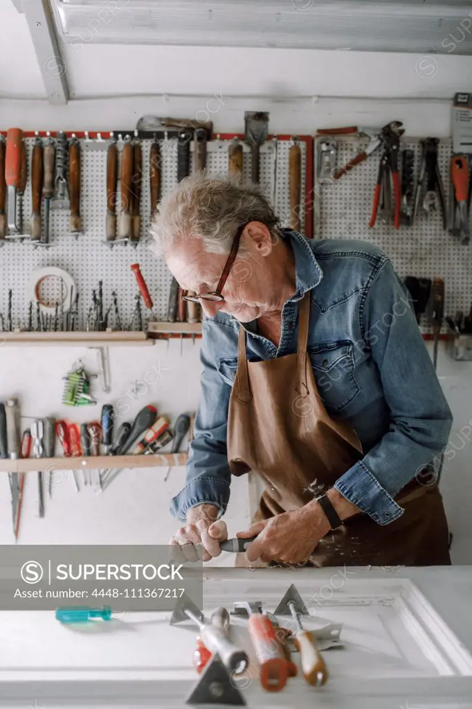 Senior male entrepreneur scraping window frame on workbench at store workshop