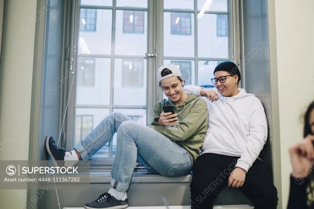 Smiling male teenage students using phone while sitting on window in classroom