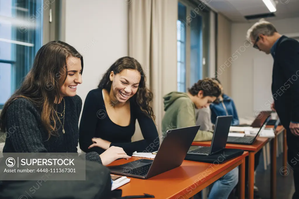 Cheerful female high school students using laptop at desk in classroom