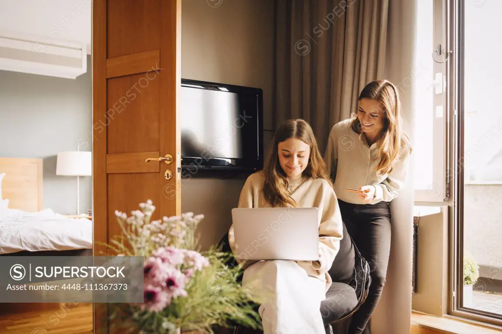 Happy friends shopping online with credit card through laptop in hotel room during vacations