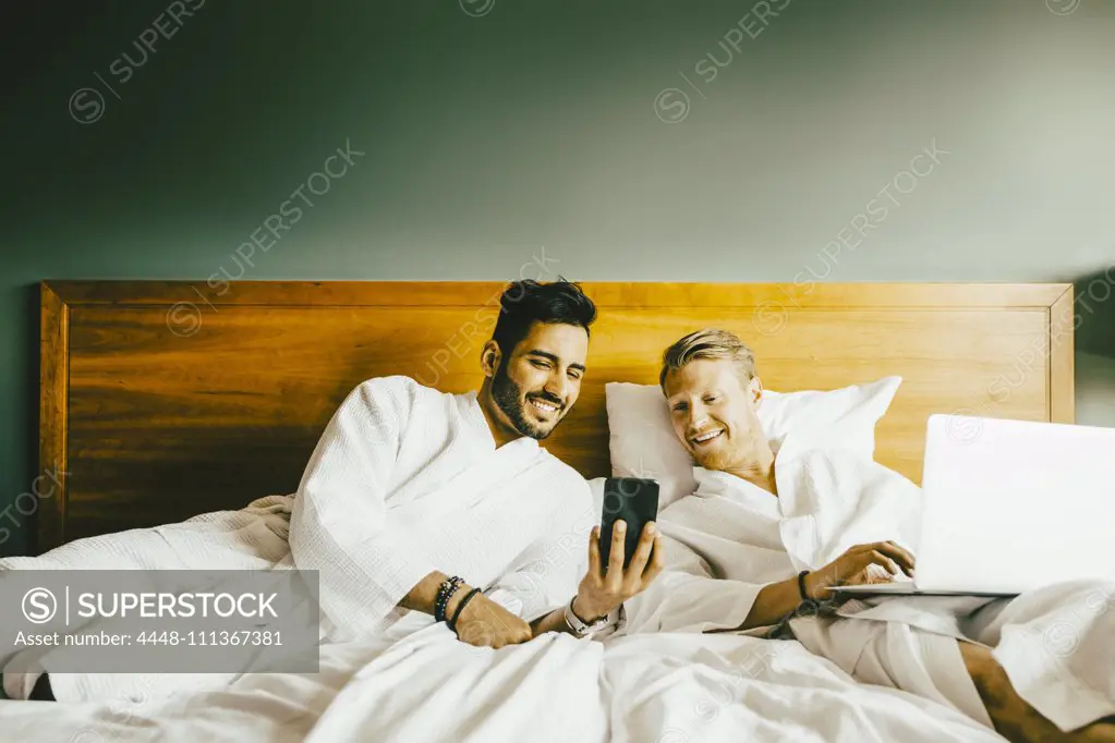 Smiling man showing smart phone to boyfriend using laptop while leaning in bed at hotel