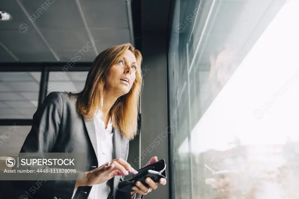 Thoughtful businesswoman with mobile phone looking through window in office