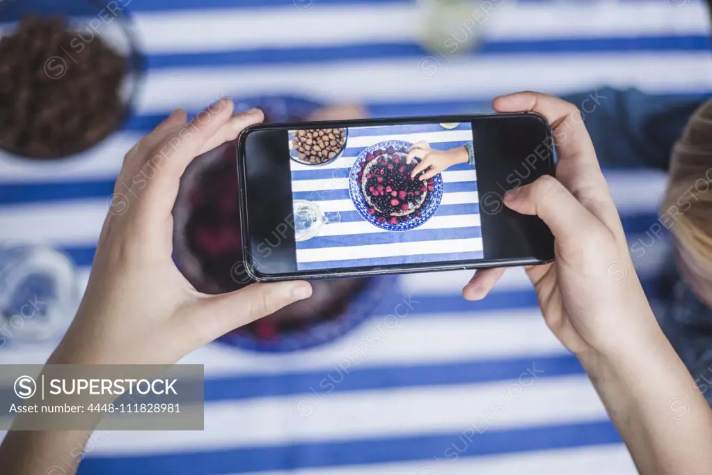 Cropped hand of girl with smart phone photographing boy having dessert at table