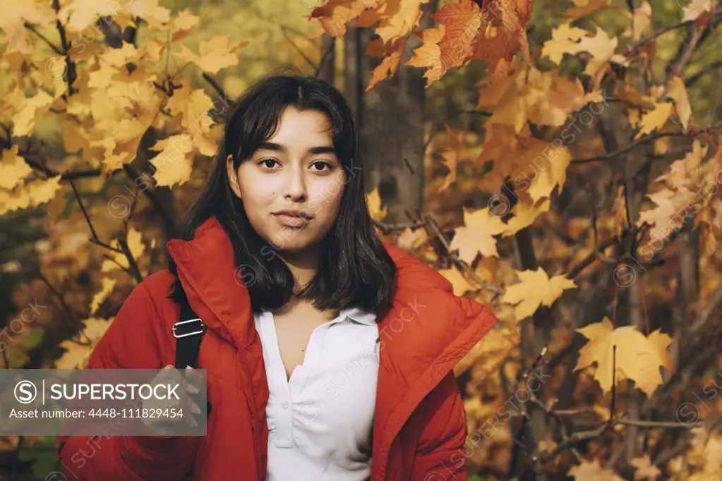 Portrait of confident teenage girl wearing red jacket standing against maple trees