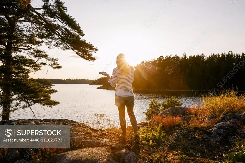 Rear view of woman holding mobile phone while standing on lakeshore at sunset