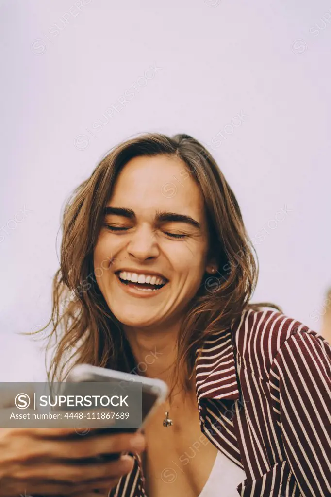 Cheerful young woman looking at smart phone held by friend at terrace