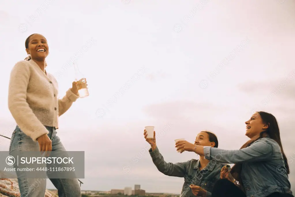 Happy woman giving drink to female friends against sky during picnic