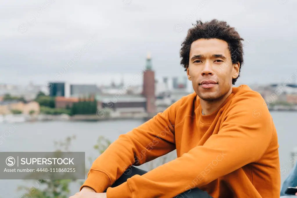 Portrait of young man sitting by cityscape against sky