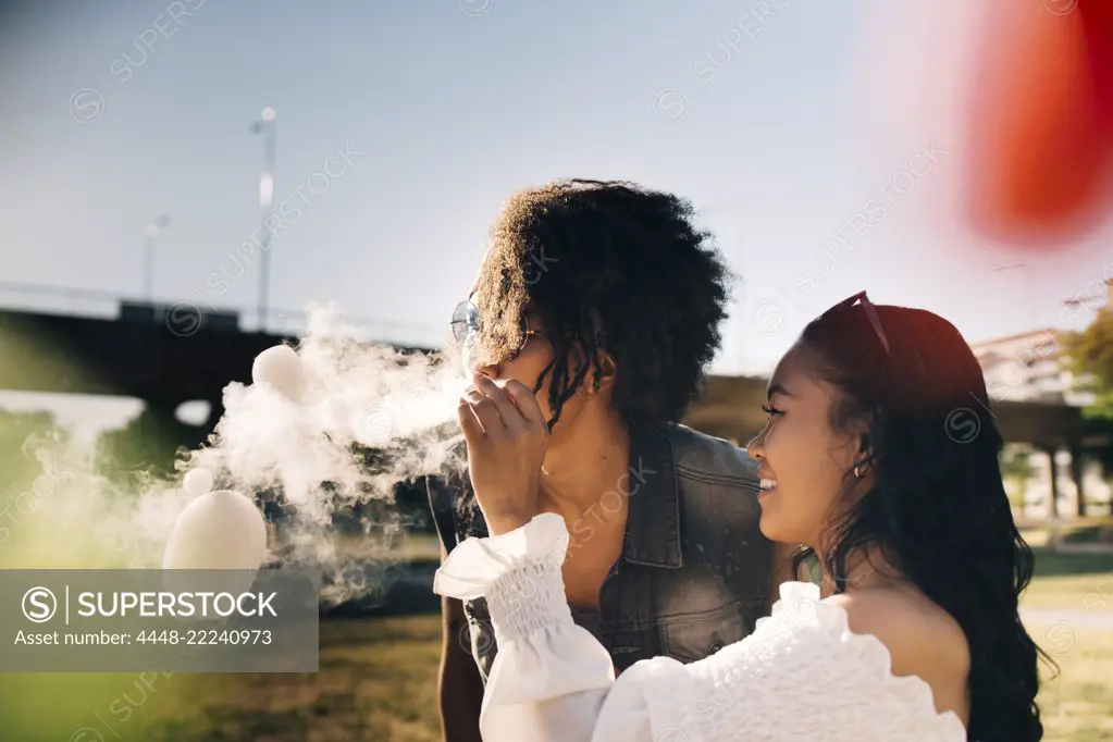 Playful woman looking at man exhaling smoke bubbles in music festival