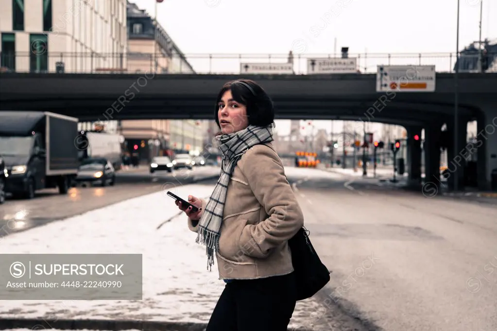 Side view of young woman holding mobile phone while crossing road in city during winter