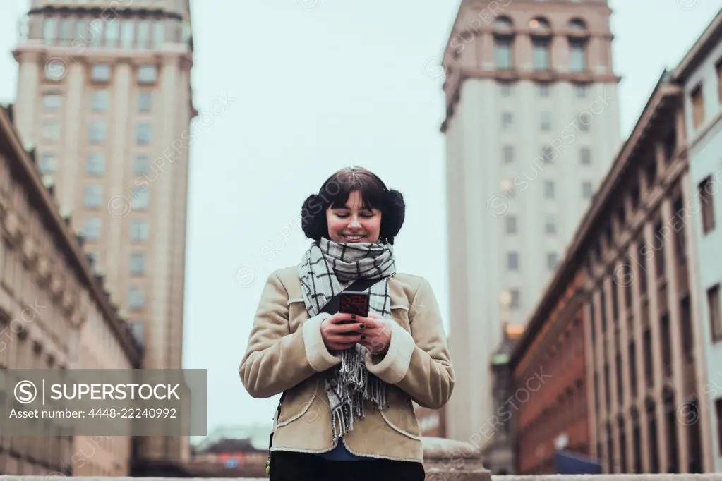 Smiling young woman wearing warm clothing while using mobile phone against buildings in city