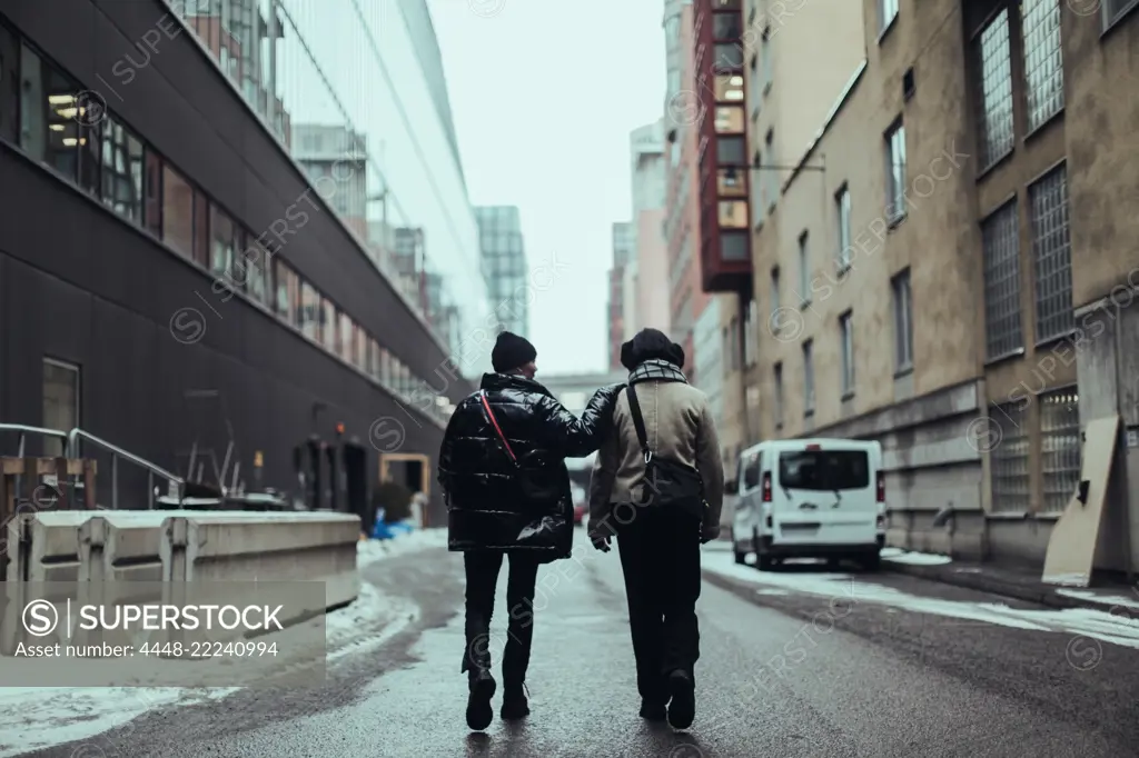 Full length rear view of female friends wearing warm clothing while walking on wet street in city by buildings