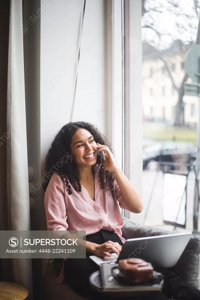 Smiling young female entrepreneur talking through smart phone sitting on window sill in office