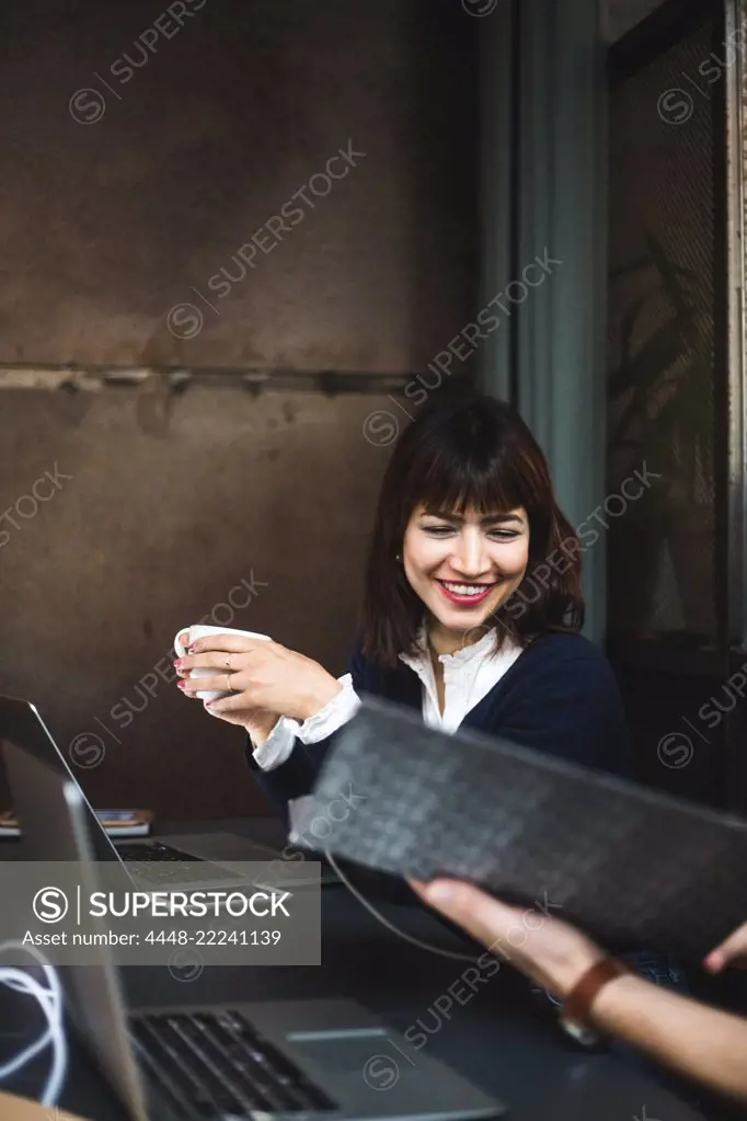 Smiling female entrepreneur holding coffee cup discussing with colleague over document at desk in office