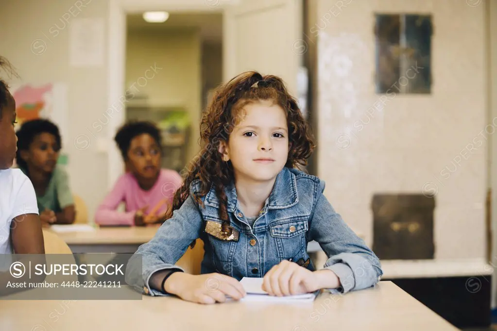 Portrait of smiling schoolgirl sitting with friends at desk in classroom