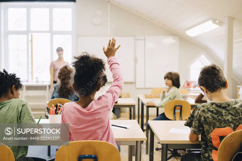 Rear view of boy raising hand while answering in class at elementary school