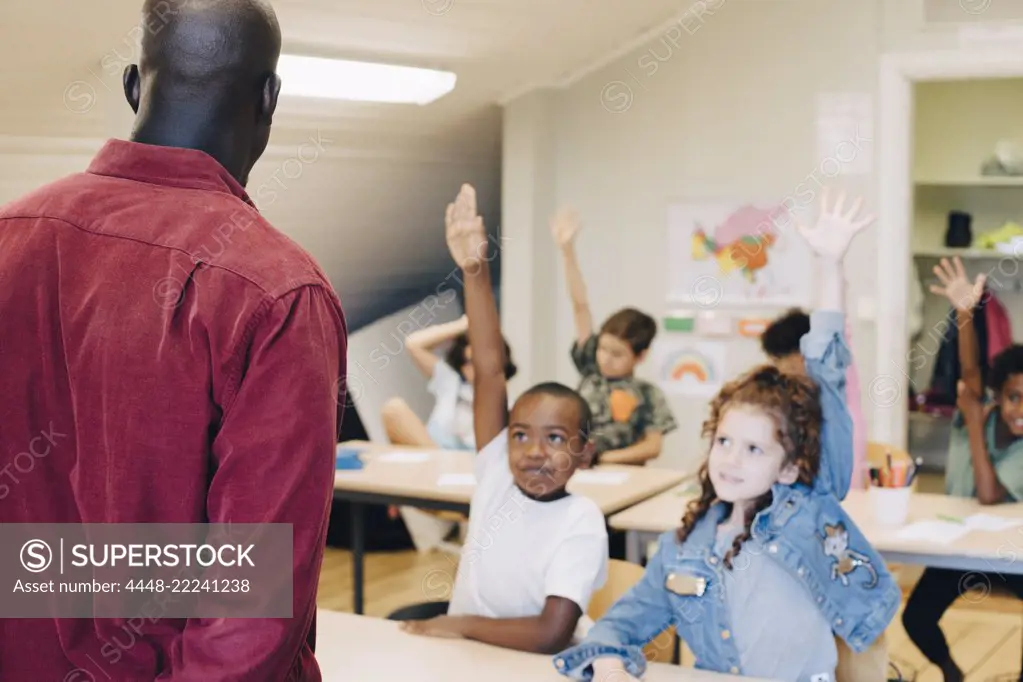 Students raising hand while looking at teacher in classroom at school