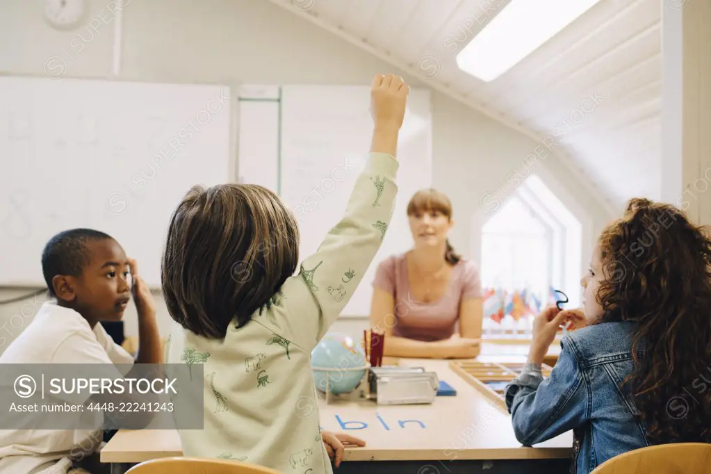 Schoolboy raising hand while answering teacher in classroom at school