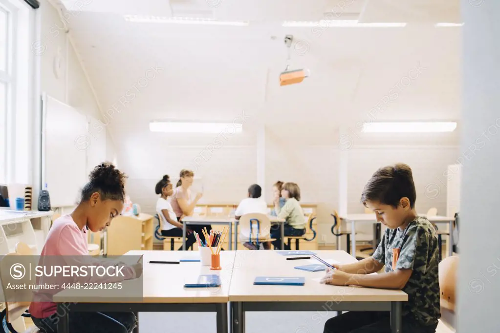 Boy and girl writing at desk while friends learning with student in background