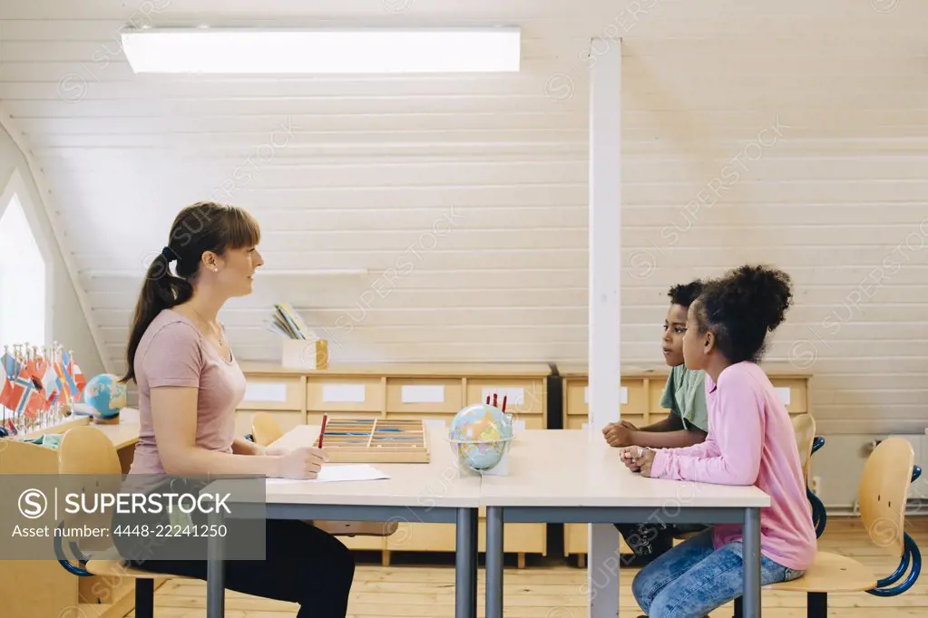 Teacher talking to school boys while sitting at desk in classroom