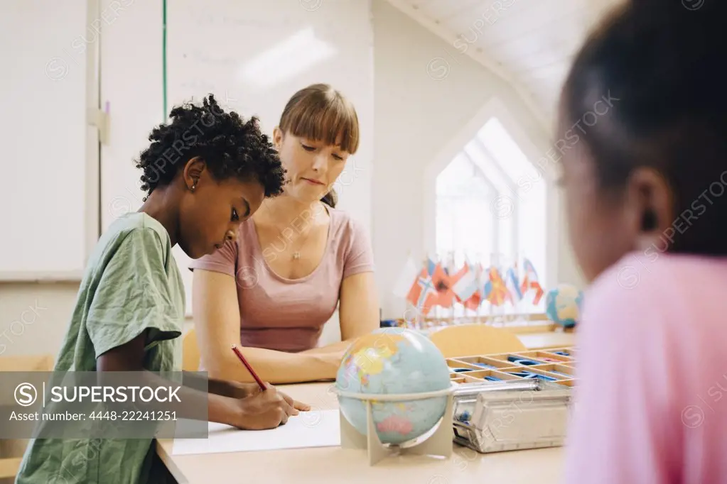 Teacher looking at boy writing on paper at table in classroom